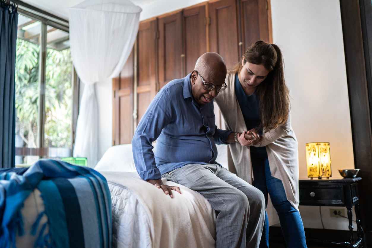 Home caregiver helping a senior man standing up at home; benefits of at-home senior care