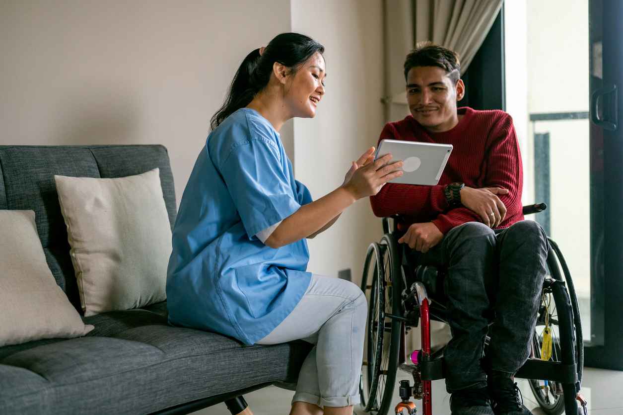 Professional Asian female healthcare worker teaching Mature Asian disabled Malay man in wheelchair how to online by using digital table at living room.