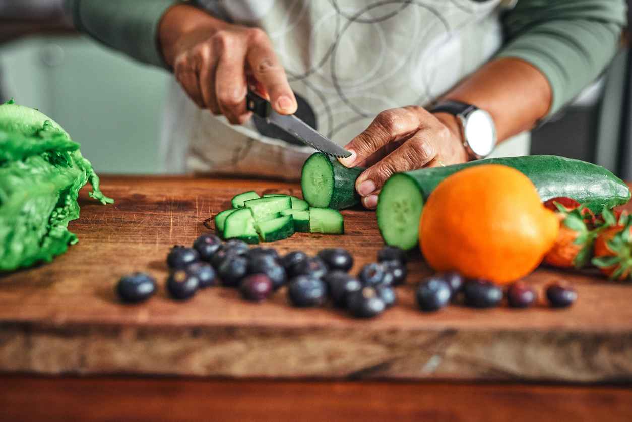 Closeup of a man slicing a cucumber on a cutting board with olives and an orange nearby