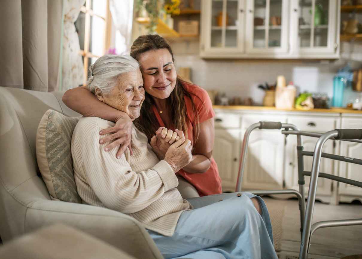 daughter assisting Senior Woman with Walker at home, hugging after senior has sat down