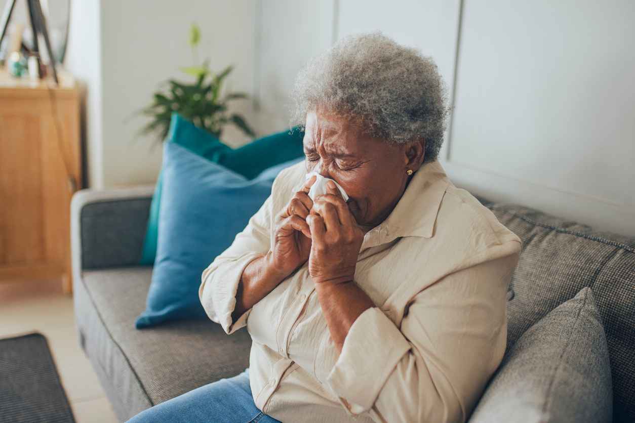 Senior woman with common cold sneezing into tissue on sofa
